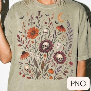 K&ouml;nnte beinhalten: Ein salbeigr&uuml;nes T-Shirt mit einem grafischen Druck eines botanischen Arrangements aus orangefarbenen und violetten Blumen, menschlichen Sch&auml;deln, kleinen geisterhaften Figuren, Flederm&auml;usen und einer Mondsichel auf der Vorderseite.