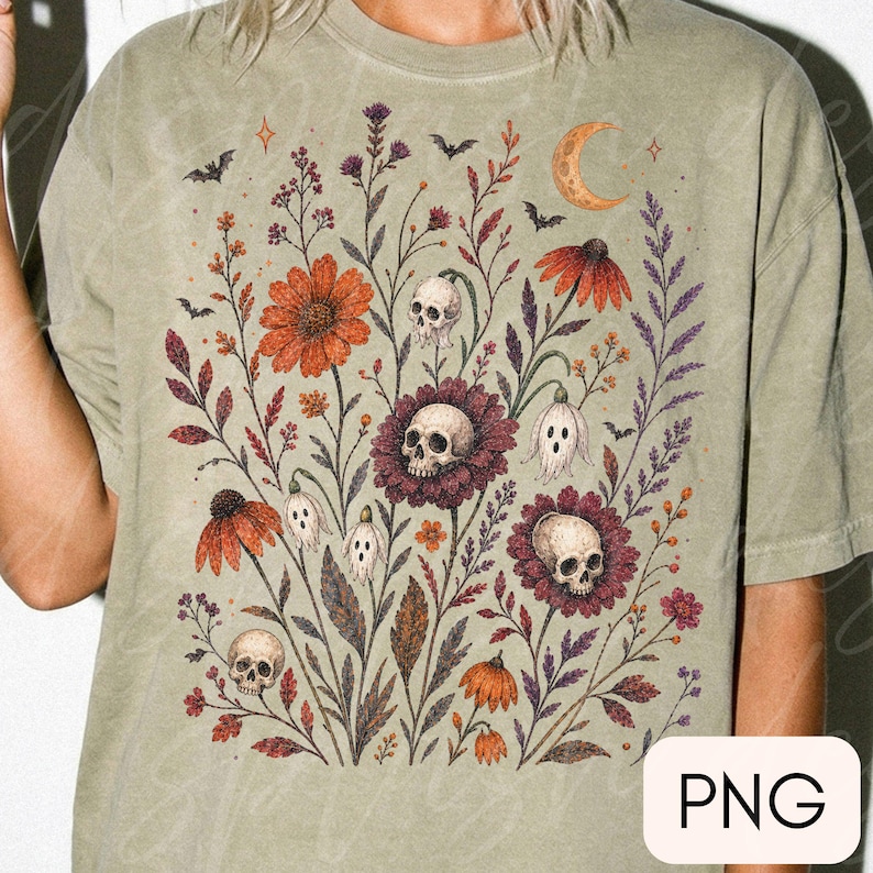 K&ouml;nnte beinhalten: Ein salbeigr&uuml;nes T-Shirt mit einem grafischen Druck eines botanischen Arrangements aus orangefarbenen und violetten Blumen, menschlichen Sch&auml;deln, kleinen geisterhaften Figuren, Flederm&auml;usen und einer Mondsichel auf der Vorderseite.