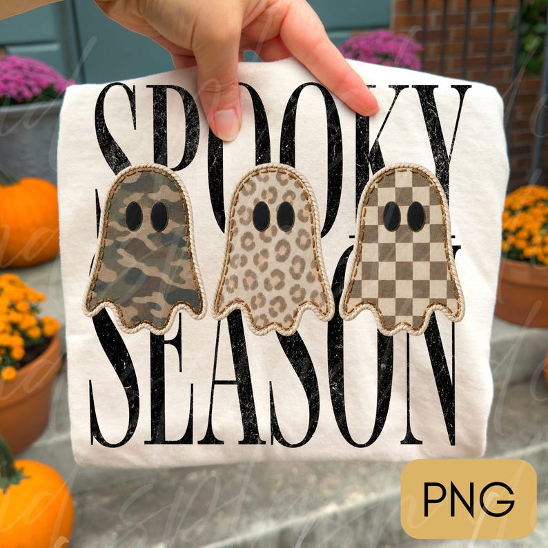 May include: A white t-shirt featuring the black distressed text SPOOKY SEASON. Three ghost shapes are centered, each with a different pattern: one with camouflage, one with leopard print, and one with a brown and white checkerboard pattern.