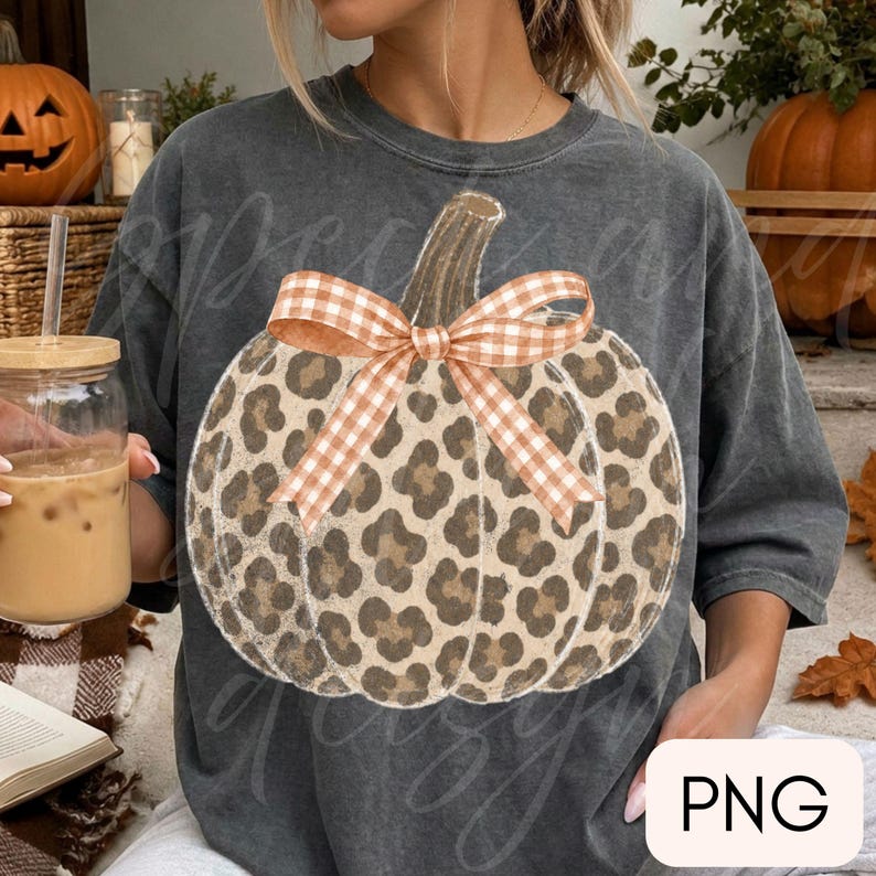 May include: A charcoal grey short-sleeve t-shirt featuring a large graphic of a pumpkin with a brown leopard print pattern. The pumpkin is decorated with a light orange and white gingham patterned bow tied at the top of the stem.