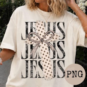 May include: A cream-coloured short-sleeve t-shirt featuring the word JESUS printed in large black distressed letters. Overlaid on the text is a beige cross with brown polka dots and a decorative bow with a glittery outline centred on the cross.