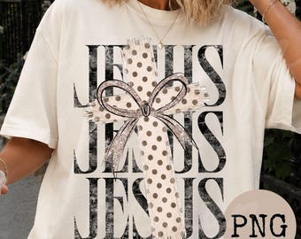 May include: A cream-colored short-sleeve t-shirt featuring the word JESUS printed in large black distressed letters. Overlaid on the text is a beige cross with brown polka dots and a decorative bow with a glittery outline centered on the cross.