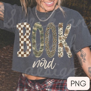 Op de afbeelding: Een antracietgrijs t-shirt met korte mouwen met het woord BOOK in grote letters met gevarieerde patronen: bruin en wit schaakbord, groen camouflage, bruine luipaardprint en een bloemmotief, met het woord nerd eronder geschreven.