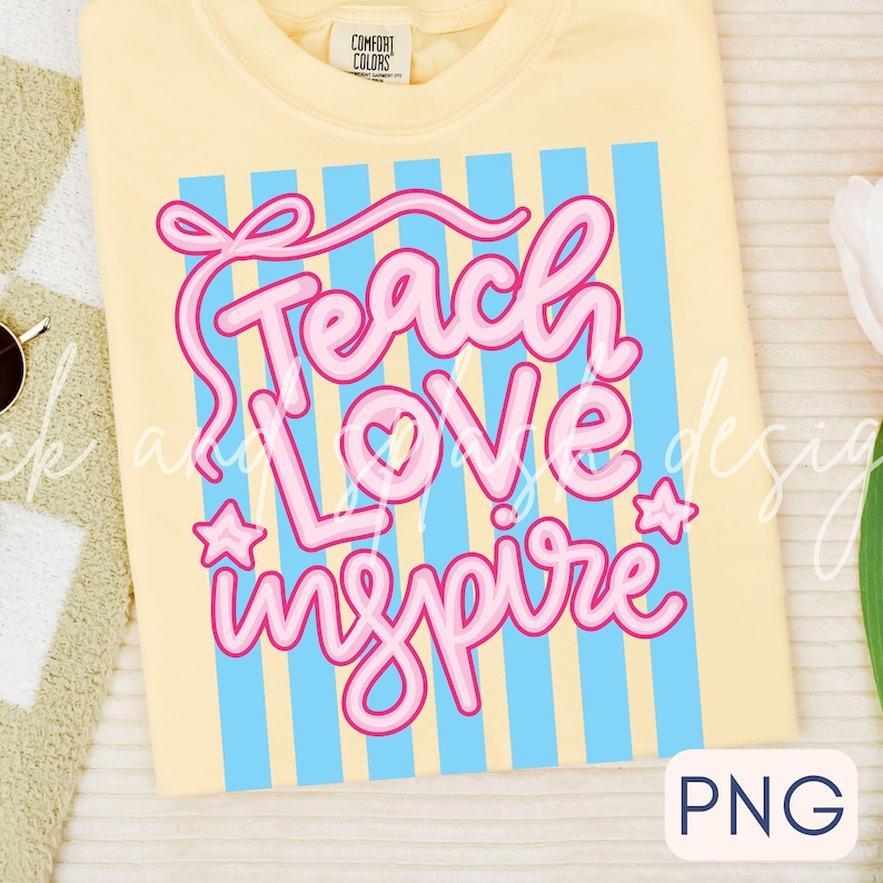 May include: A light yellow t-shirt with the words "Teach Love Inspire" in pink script, accented with blue vertical stripes. The shirt has a Comfort Colors tag.
