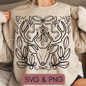 Puede incluir: Sudadera beige con un dibujo lineal negro de una cabeza de ciervo con cuernos, muérdago y hojas. El texto "SVG & PNG" está en un rectángulo granate en la parte inferior. El diseño está centrado en la parte delantera.
