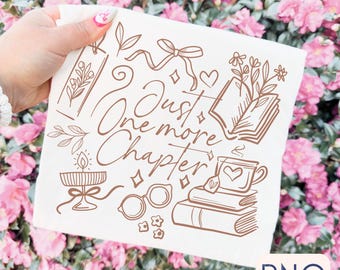 May include: A cream-coloured square textile featuring a brown line drawing of books, a teacup, glasses, and the phrase "Just One More Chapter." The design includes floral and ribbon accents. The background is a blur of pink flowers.
