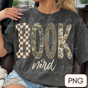 Op de afbeelding: Een antracietgrijs T-shirt met korte mouwen en het woord BOOK in grote letters met verschillende patronen: bruin dambord, groen camouflage, luipaardprint en een bloemendesign. Het woord nerd staat eronder in witte schrijfletters.