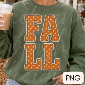 May include: An olive green crewneck sweatshirt featuring the word FALL in large, block-style letters. The letters are orange with a white polka dot pattern and a thick, rope-like white border that mimics a stitched or embroidered appearance.