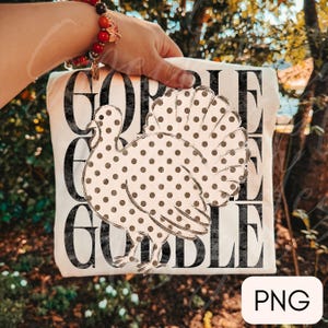 May include: A white t-shirt featuring a large, central graphic of a turkey with a brown polka dot pattern. Behind the turkey, the word GOBBLE is printed three times in a distressed, black, serif font, stacked vertically across the shirt front.