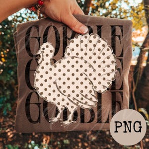 May include: A brown t-shirt featuring a large white turkey graphic covered in dark brown polka dots. The background of the design displays the word GOBBLE repeated three times in a bold, dark brown, distressed sans-serif font.