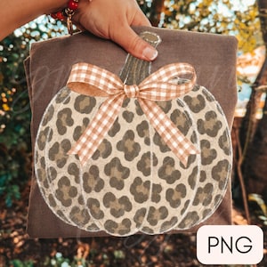 May include: A brown t-shirt featuring a large pumpkin graphic with a tan and brown leopard print pattern. The pumpkin is adorned with a tan and white gingham check bow tied at the stem. The design is centred on the front of the garment.