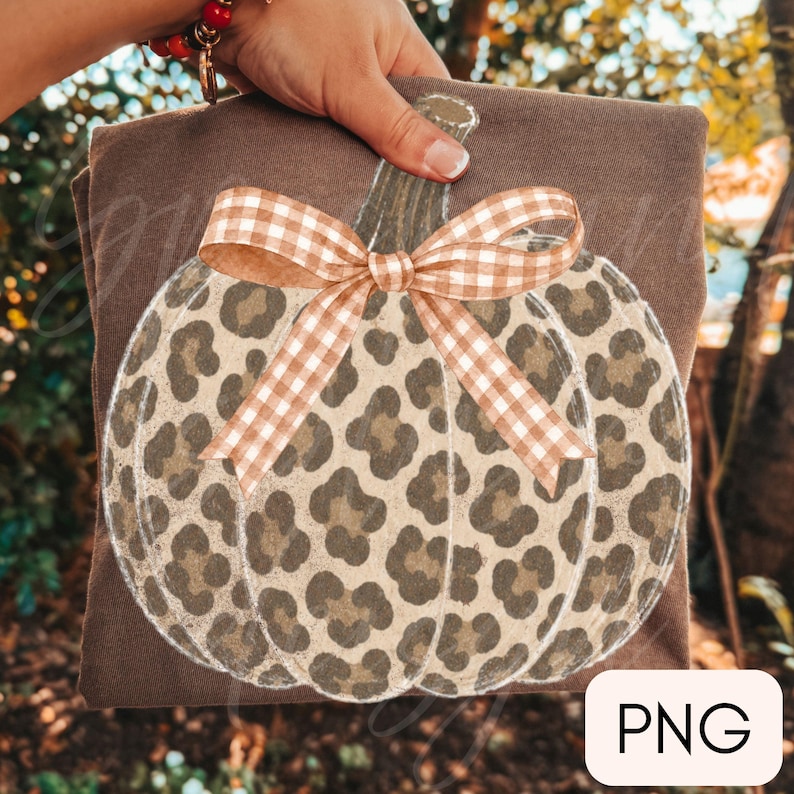 May include: A brown t-shirt featuring a large pumpkin graphic with a tan and brown leopard print pattern. The pumpkin is adorned with a tan and white gingham check bow tied at the stem. The design is centred on the front of the garment.