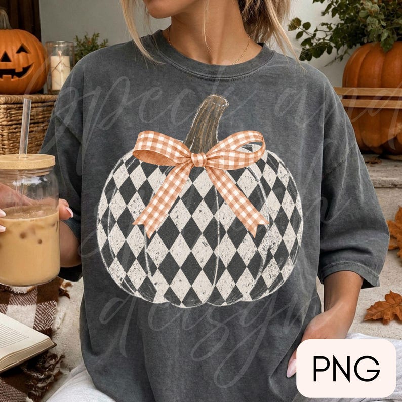 May include: A charcoal gray short-sleeve t-shirt featuring a large graphic of a pumpkin with a black and white diamond harlequin pattern. The pumpkin is adorned with a light orange and white gingham check bow tied around its stem.