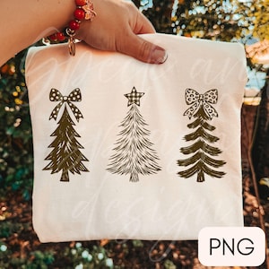 May include: A cream-colored t-shirt featuring a graphic print of three dark green Christmas trees. The first tree has a polka dot bow, the second has a plaid star topper, and the third has a leopard print bow. The design is centered on the fabric.