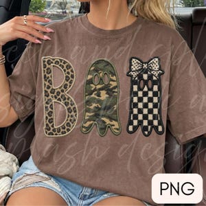 May include: A brown short-sleeve t-shirt featuring the word BOO in large letters. The B has a leopard print, the O is a ghost with a camouflage pattern, and the second O is a ghost with a black and white checkerboard pattern and a bow.