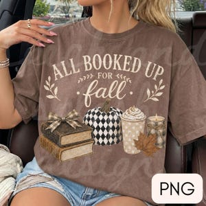 May include: A brown short-sleeve t-shirt featuring the text ALL BOOKED UP FOR fall in white and cream lettering. The graphic includes a stack of two books with a plaid bow, a black and white checkered pumpkin, a coffee cup, and a candle.