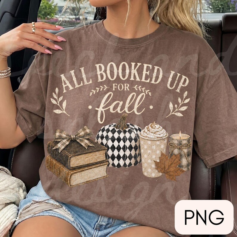 May include: A brown short-sleeve t-shirt featuring the text ALL BOOKED UP FOR fall in white and cream lettering. The graphic includes a stack of two books with a plaid bow, a black and white checkered pumpkin, a coffee cup, and a candle.