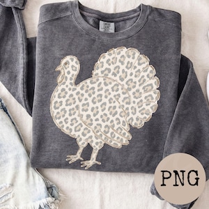 May include: A charcoal gray crewneck sweatshirt featuring a graphic of a turkey with a beige and brown leopard print pattern. The turkey design has a thin gold outline. The garment has a label at the inner neckline that reads Comfort Colors.
