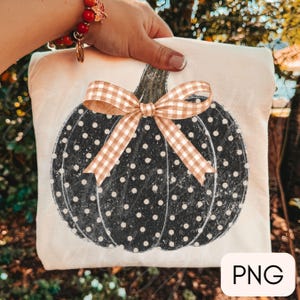 May include: A cream-colored fabric item featuring a black pumpkin graphic with white polka dots and a tan and white gingham bow tied at the top. The pumpkin design has a distressed texture and is centered on the light-colored textile surface.