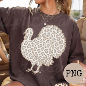 May include: A dark brown short-sleeve t-shirt featuring a large graphic of a turkey with a beige and grey leopard print pattern. The turkey graphic has a thin gold outline. The shirt has a crew neckline and a relaxed, loose-fitting silhouette.
