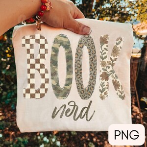 Op de afbeelding: Een cr&egrave;mekleurig t-shirt met het woord BOOK in grote blokletters, elk met een ander patroon: bruin en wit schaakbord, groen camouflage, bruine luipaardprint en een beige bloemmotief, met het woord nerd in cursief schrift eronder.