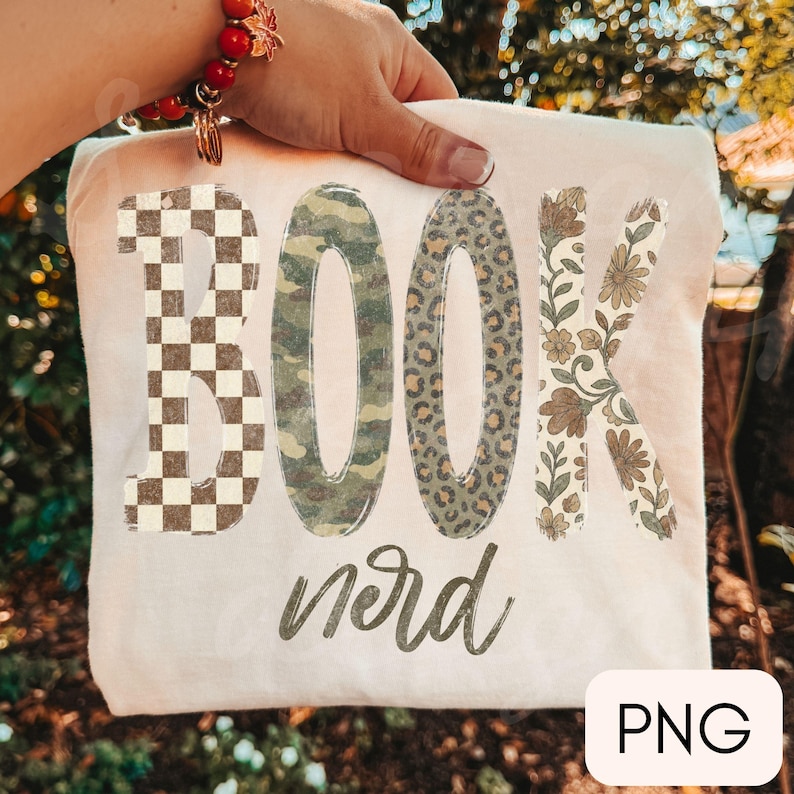 Op de afbeelding: Een cr&egrave;mekleurig t-shirt met het woord BOOK in grote blokletters, elk met een ander patroon: bruin en wit schaakbord, groen camouflage, bruine luipaardprint en een beige bloemmotief, met het woord nerd in cursief schrift eronder.