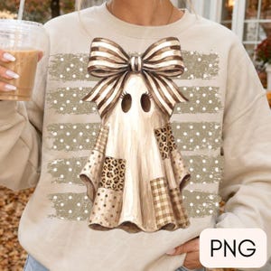 May include: A beige crewneck sweatshirt featuring a central graphic of a ghost wearing a large striped bow. The ghost has patches with leopard print, plaid, and polka dot patterns. Behind the ghost are horizontal beige brush strokes with white dots.