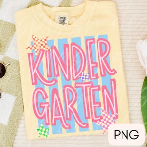 May include: A pale yellow t-shirt featuring the text KINDERGARTEN in large pink letters. The design includes vertical light blue stripes and several small, multi-colored checkered stars scattered across the front. The shirt has a Comfort Colors label.