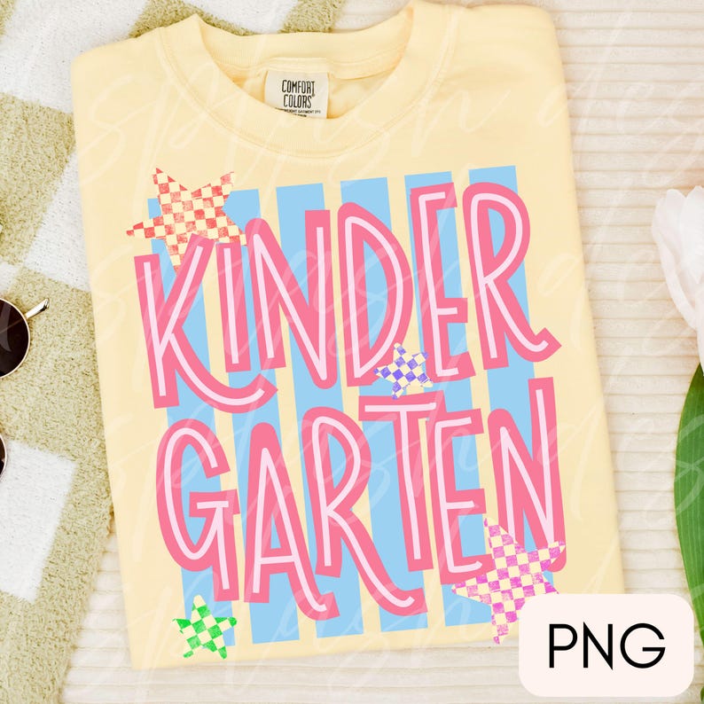 May include: A pale yellow t-shirt featuring the text KINDERGARTEN in large pink letters. The design includes vertical light blue stripes and several small, multi-colored checkered stars scattered across the front. The shirt has a Comfort Colors label.