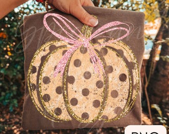 May include: A brown t-shirt featuring a graphic of a tan pumpkin with brown polka dots. The pumpkin is outlined in gold glitter and topped with a large, pink glitter bow. The design has a textured, distressed appearance on the fabric.