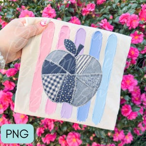 May include: A cream-colored t-shirt featuring a central apple graphic composed of various blue denim fabric patches with frayed edges. Behind the apple are five vertical brushstroke stripes in shades of pink, lavender, and light blue.