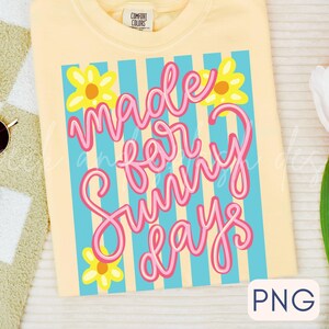 May include: A light yellow t-shirt with the words "Made for Sunny Days" in pink script, set against a background of blue stripes and yellow flowers. The shirt is a Comfort Colors brand.