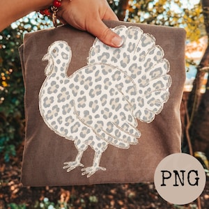 May include: A brown t-shirt featuring a graphic of a turkey with a light gray and cream leopard print pattern. The turkey design is outlined in a thin gold color. The shirt is held up against a blurred outdoor background with trees.