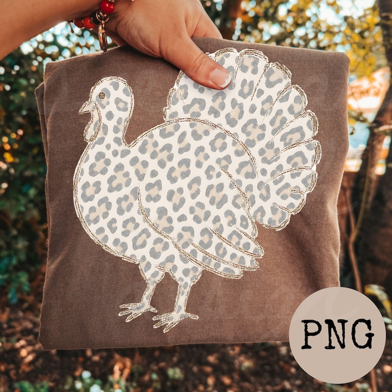 May include: A brown t-shirt featuring a graphic of a turkey with a light gray and cream leopard print pattern. The turkey design is outlined in a thin gold color. The shirt is held up against a blurred outdoor background with trees.