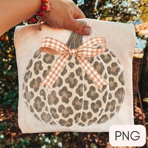 May include: A cream-colored fabric item featuring a central graphic of a pumpkin with a tan and brown leopard print pattern. The pumpkin is adorned with a tan and white gingham check bow tied at the top stem. The letters PNG appear in a box.