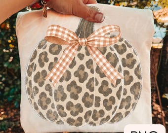 May include: A cream-colored fabric item featuring a central graphic of a pumpkin with a tan and brown leopard print pattern. The pumpkin is adorned with a tan and white gingham check bow tied at the top stem. The letters PNG appear in a box.