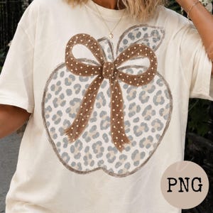 May include: A cream-coloured short-sleeve t-shirt featuring a large graphic of an apple with a grey and tan leopard print pattern. The apple is adorned with a brown bow that has white polka dots. The text PNG is visible in the bottom right corner.