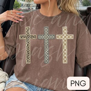 May include: A brown short-sleeve t-shirt featuring three vertical cross graphics across the chest. The left cross has a leopard print pattern, the middle cross has a white polka dot pattern on a gray background, and the right cross has a checkerboard pattern.