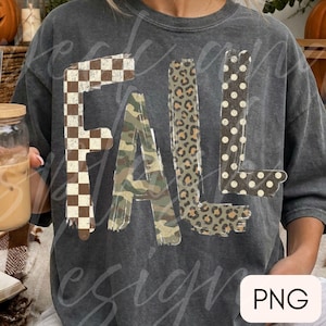 May include: A charcoal gray short-sleeve t-shirt featuring the word FALL in large block letters. Each letter has a distinct pattern: the F is brown and white checkered, the A is green camouflage, the L is leopard print, and the L is brown polka dot.