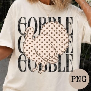 May include: A cream-colored t-shirt featuring a large graphic of a turkey with brown polka dots. Behind the turkey, the word GOBBLE is printed three times in a distressed black font, stacked vertically to create a bold, layered visual effect.