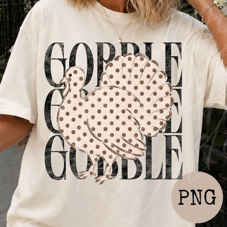 May include: A cream-colored t-shirt featuring a large graphic of a turkey with brown polka dots. Behind the turkey, the word GOBBLE is printed three times in a distressed black font, stacked vertically to create a bold, layered visual effect.