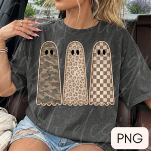 May include: A charcoal gray short-sleeve t-shirt featuring a graphic of three ghosts arranged side-by-side. The first ghost has a camouflage pattern, the second has a leopard print, and the third has a brown and white checkered pattern.