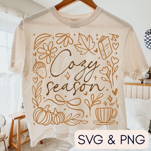 Pu&ograve; includere: T-shirt color crema con un design grafico marrone. Il design presenta le parole "Cozy Season" circondate da illustrazioni a tema autunnale, tra cui zucche, foglie, fiori e una tazza da t&egrave;. Il testo "SVG & PNG" &egrave; in basso a destra.