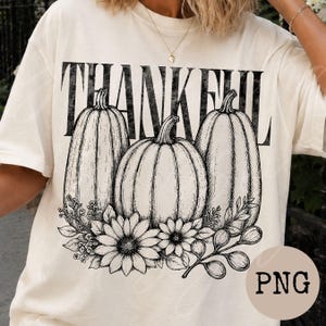 Könnte beinhalten: Ein cremefarbenes T-Shirt mit einer schwarzen Grafik im Tintenstil, die drei Kürbisse und Sonnenblumen zeigt. Das Wort THANKFUL ist in großen, abgenutzten schwarzen Großbuchstaben über den Kürbissen aufgedruckt.
