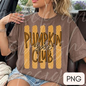 May include: A brown short-sleeved t-shirt featuring a graphic design of five vertical yellow stripes with white polka dots. The text PUMPKIN SPICE CLUB is printed in a dark brown and orange font over the stripes, with a small heart above the word spice.