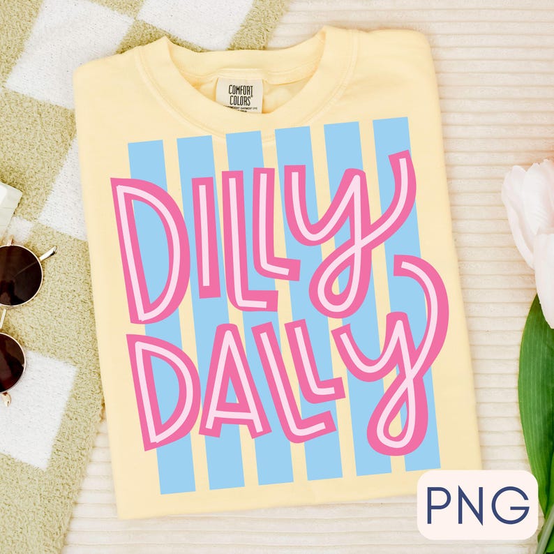 Puede incluir: Una camiseta amarilla Comfort Colors con las palabras "Dilly Dally" en rosa y blanco, sobre un fondo de rayas verticales azules. La camiseta est&aacute; doblada.