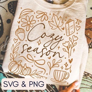 Pu&ograve; includere: Una felpa beige con un design grafico marrone. Il design include le parole "Cozy Season" circondate da illustrazioni a tema autunnale come zucche, foglie, fiori e una tazza da t&egrave;. La parte inferiore dell'immagine mostra "SVG & PNG".