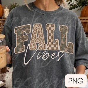 May include: A charcoal grey short-sleeve t-shirt featuring the word FALL in large block letters with varied patterns including camouflage, leopard print, and a checkerboard design. The word Vibes is written below in a white cursive script.
