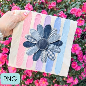 Op de afbeelding: Een wit t-shirt met verticale penseelstreken in roze, lavendel en lichtblauwe tinten. In het midden bevindt zich een grote madelief-applicatie gemaakt van diverse blauwe spijkerstof lapjes, inclusief ruitjes- en streepmotieven.