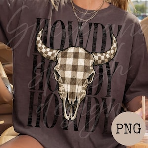 May include: A dark brown t-shirt featuring a large graphic of a bull skull with a beige and brown checkered pattern. The word HOWDY is printed in large, distressed black capital letters repeatedly in the background behind the bull skull graphic.